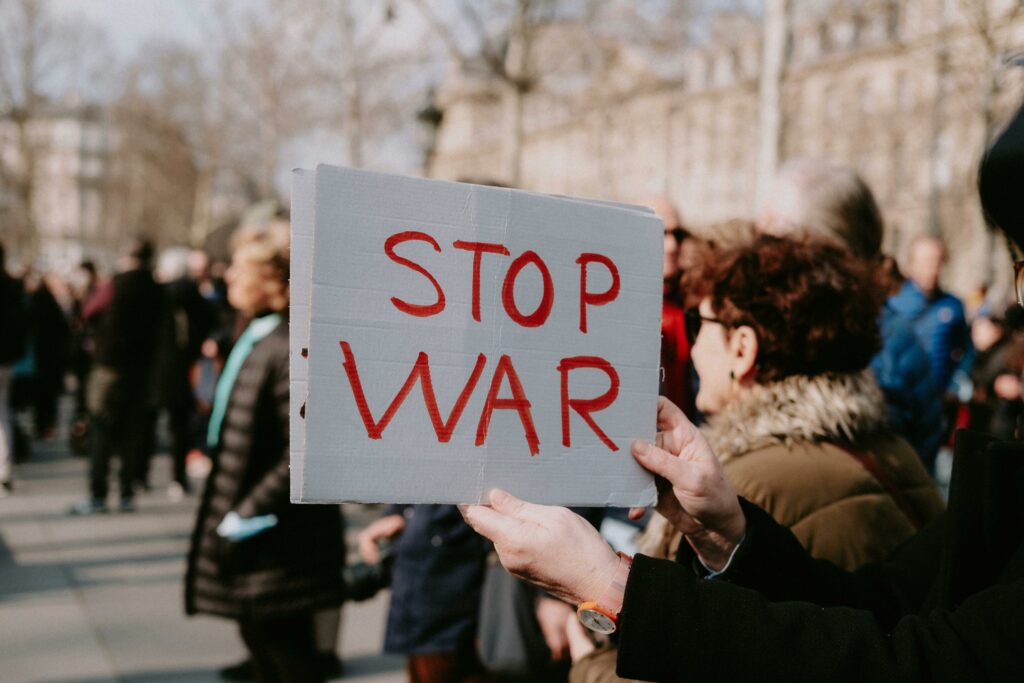 Das Bild zeigt eine Demonstration. Im Vordergrund hebt eine Person ein weißes Schild mit der roten Aufschrift "Stop War" also "Stoppt den Krieg".